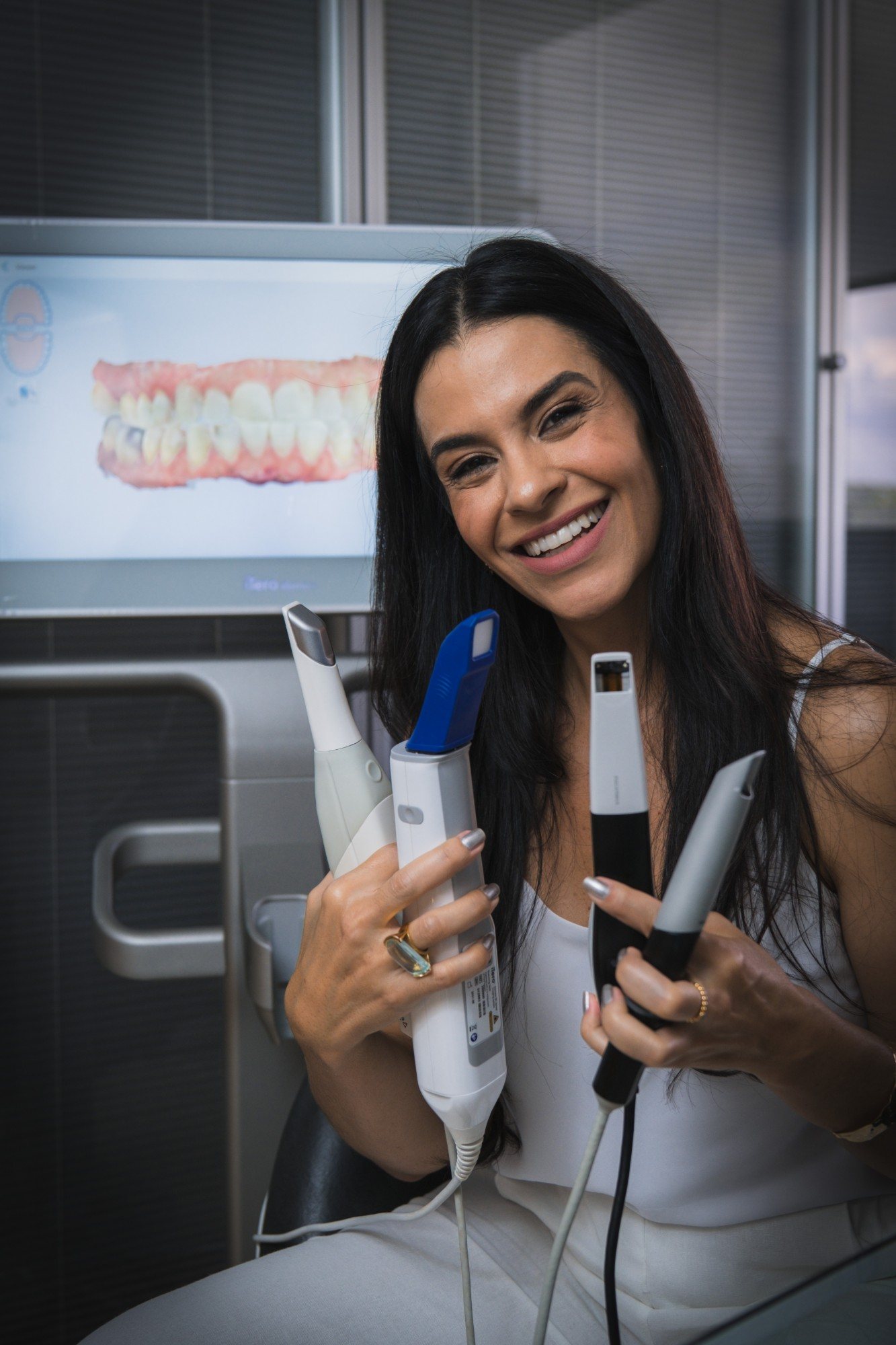 Ananda Marga segurando scanners intraorais de diferentes marcas, com um escaneamento na tela ao fundo